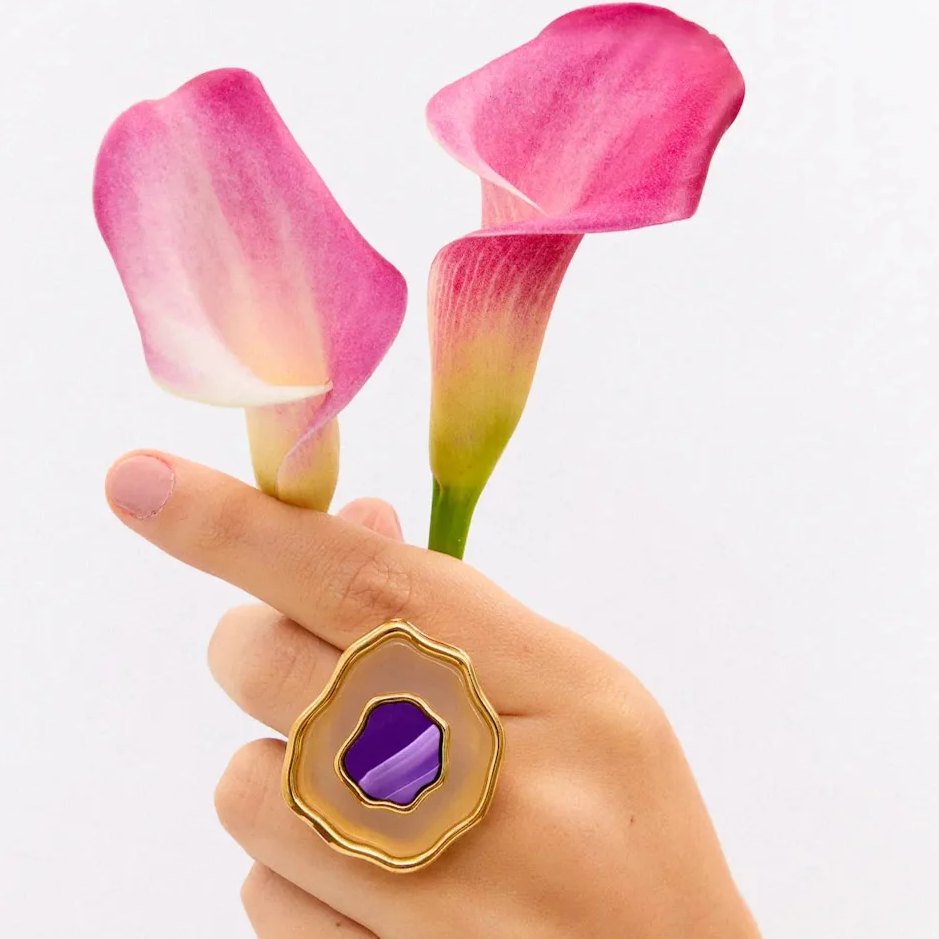 Mão segurando copos-de-leite cor-de-rosa e usando o anel Anthera com Ágata Roxa Listrada, semijoia banhada a ouro 18k, Aimer Joias