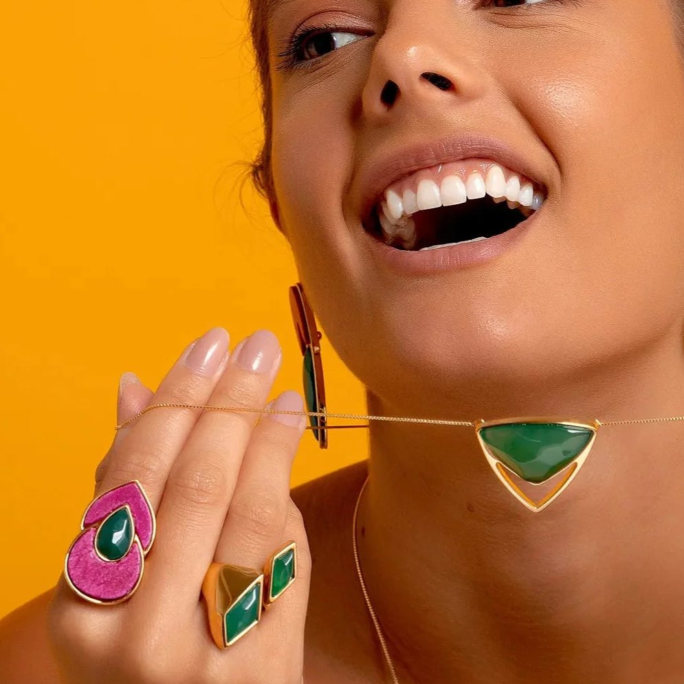 Modelo sorrindo sobre fundo amarelo, com anéis Flor de Lótus de Ágata Verde e Feldspato Rosa Pink, semijoias banhadas a ouro 18k, Aimer Joias