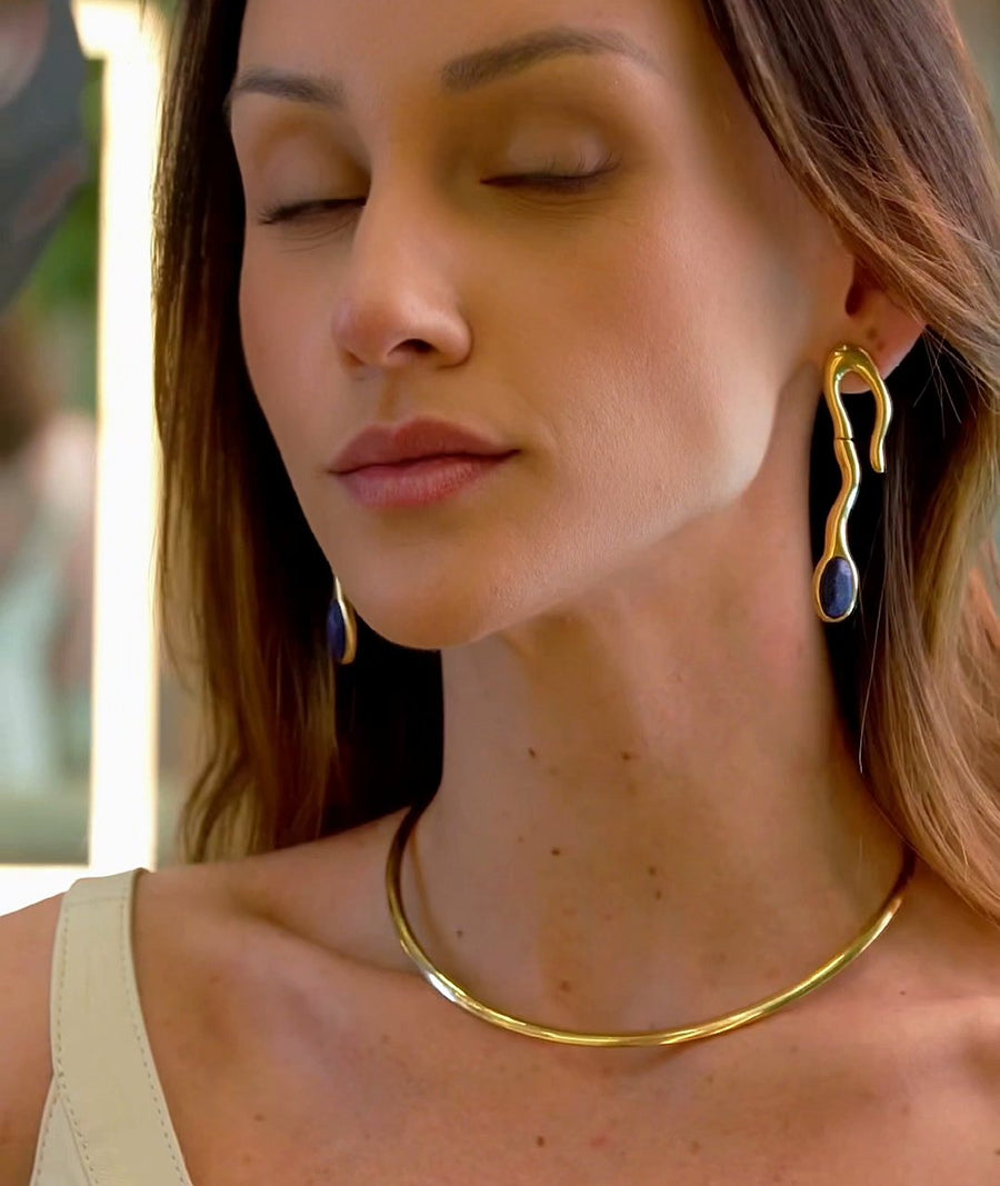 Mari de olhos fechados usando o Aro Ondulado, colar rígido banhado a ouro 18k, e o Brinco Noha